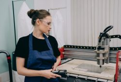 Woman in workshop using CNC machine, wearing goggles and apron, focused on industrial work.